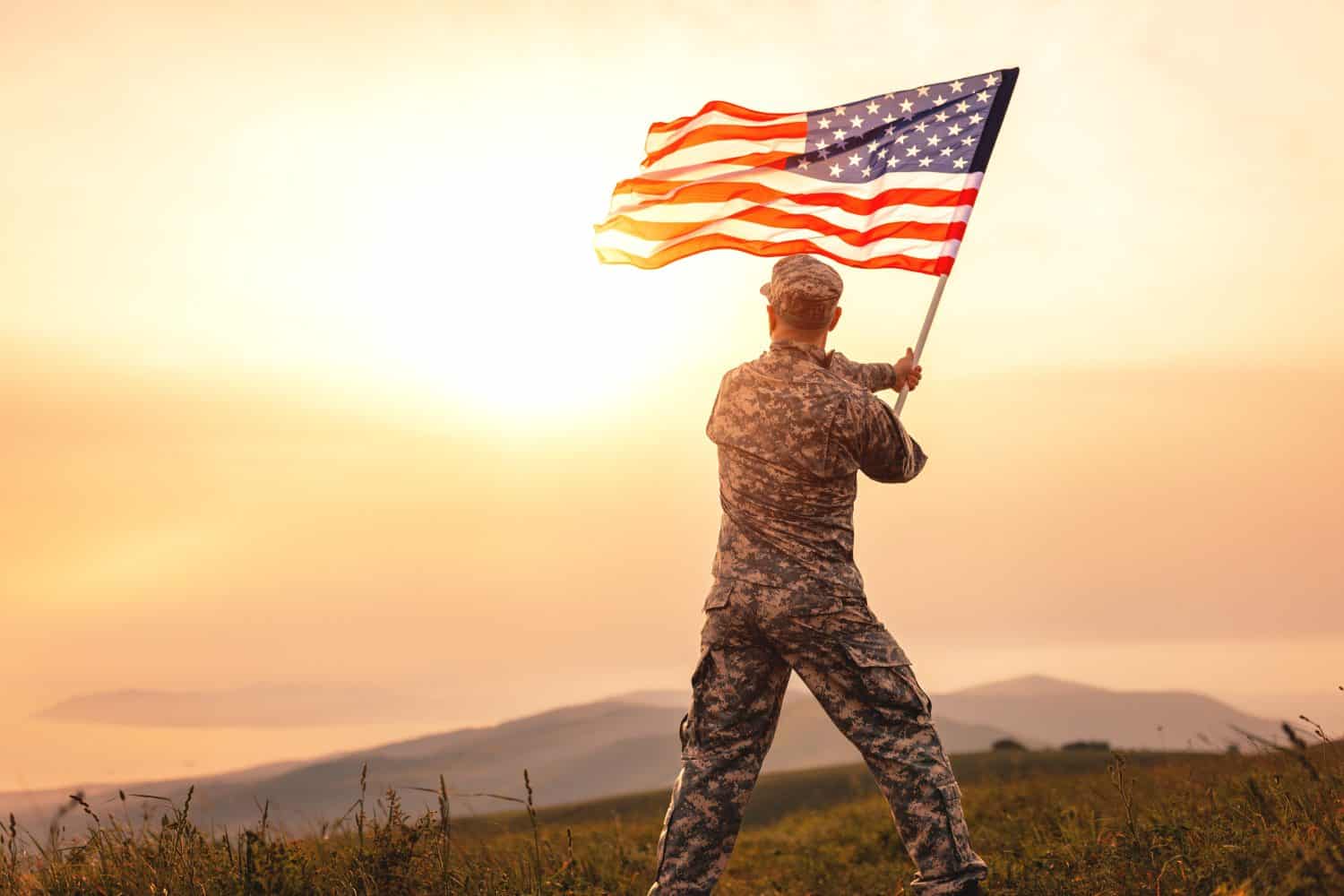 View from the back of a male soldier in the uniform of the American army waving the US flag on top of a mountain in a clearing at sunset by Evgeny Atamanenko