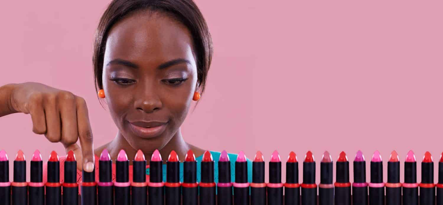 Lipstick, makeup and choice of color with black woman in studio isolated on pink background for cosmetics. Face, retail and shopping for beauty product with young customer on space for decision