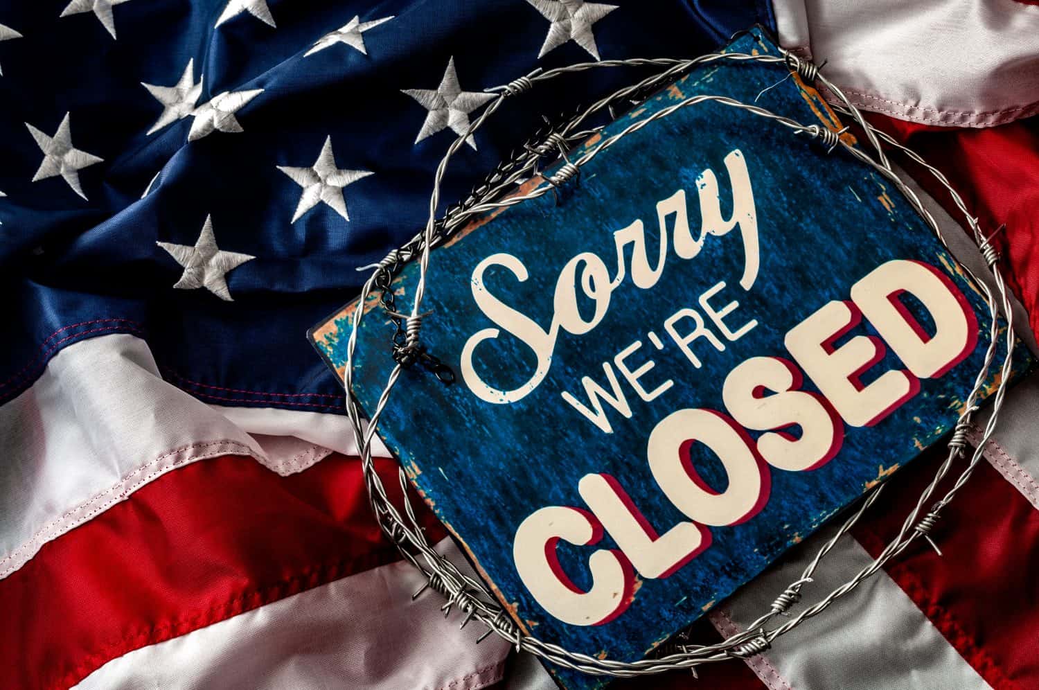 Political isolationism and economic slowdown in the United States of America concept with close up a sorry we&#039;re closed sign wrapped in barbwire on top of the american flag