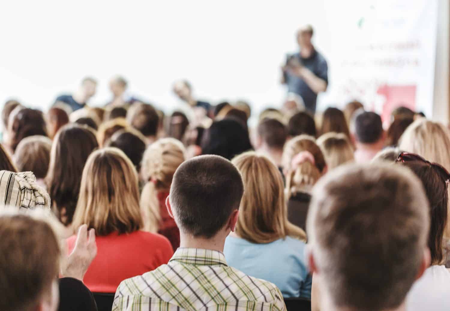 Audience in small classroom. Adult students listen to professor. Group of professionals in audience listening to speaker. Rear view sitting people, business concept.