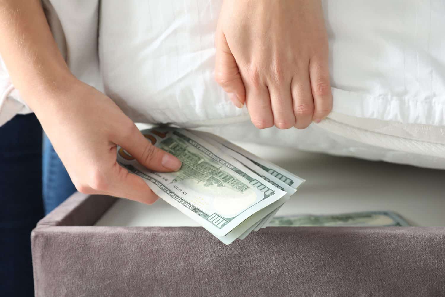 Woman hiding dollar banknotes under mattress in bedroom, closeup. Money savings