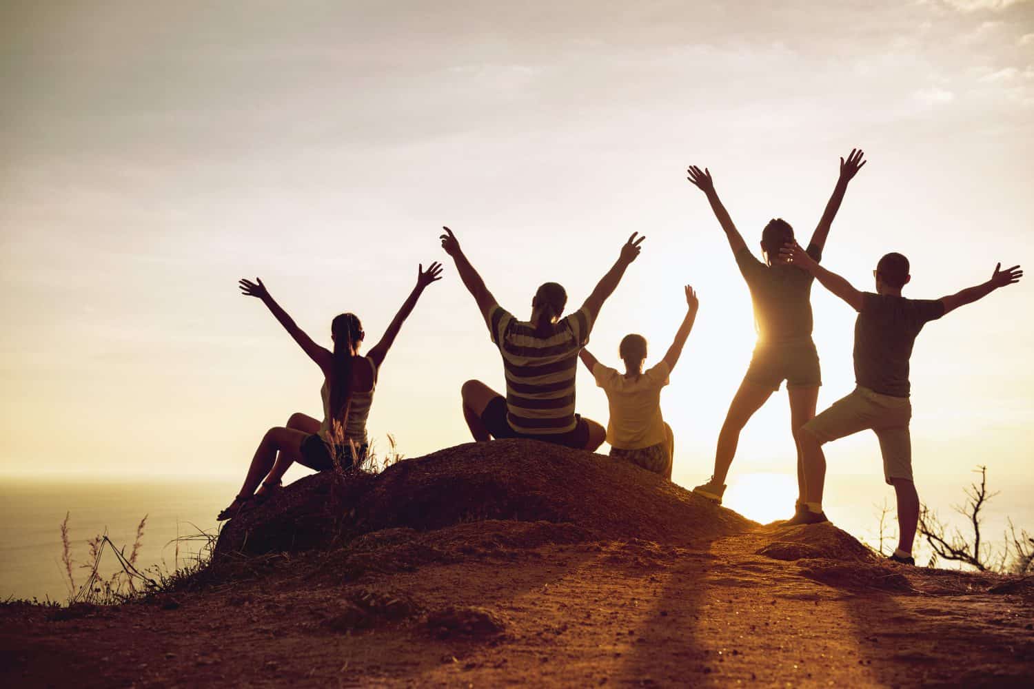 Group of happy tourists stands and sits with raised arms at sunset and having fun