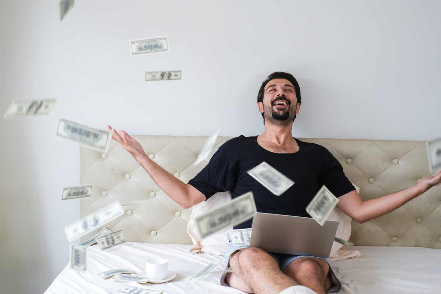 Handsome joyful young businessman sitting on bed, working from home throwing money over his head. Happy Smiling man throws the money up in the air and sitting on bed in hotel room.