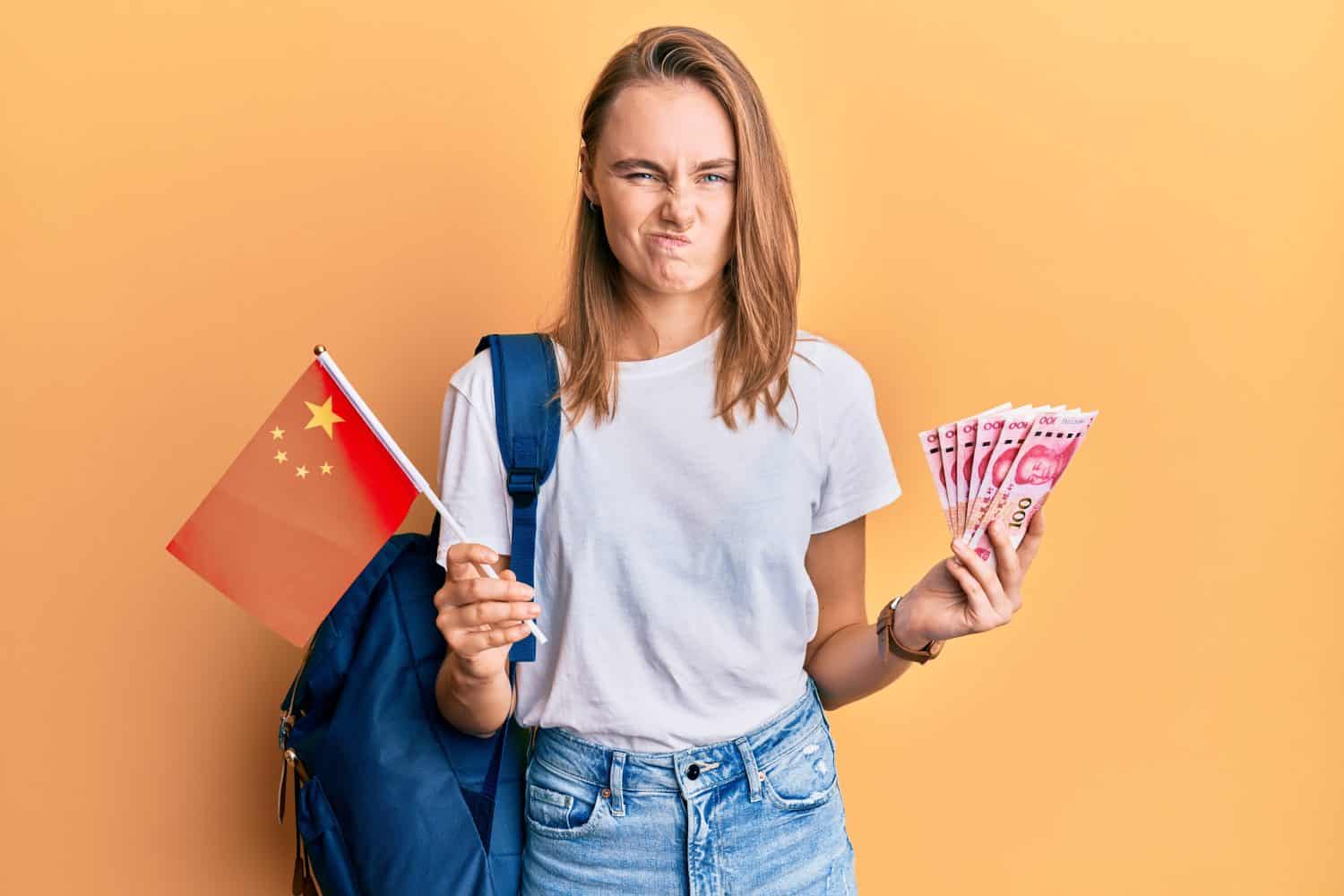 Beautiful blonde woman exchange student holding china flag and yuan banknotes skeptic and nervous, frowning upset because of problem. negative person.