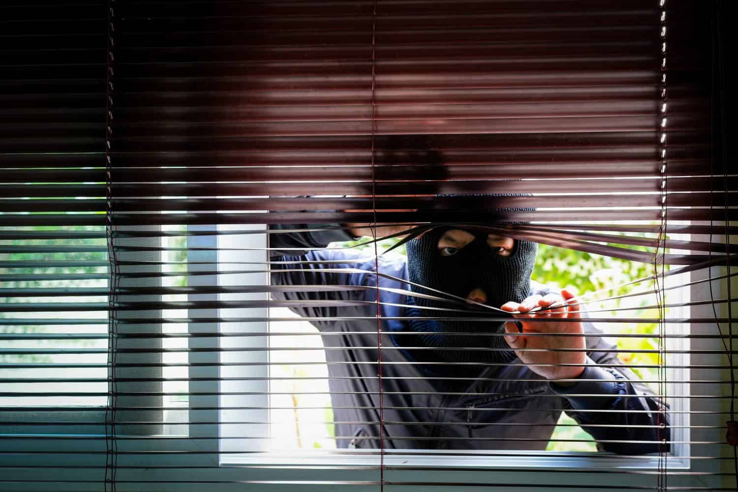 Unrecognizable Man Wearing Balaclava Face Mask Looking Through Venetian Blinds. Keep your home secure during your summer holiday Concept.