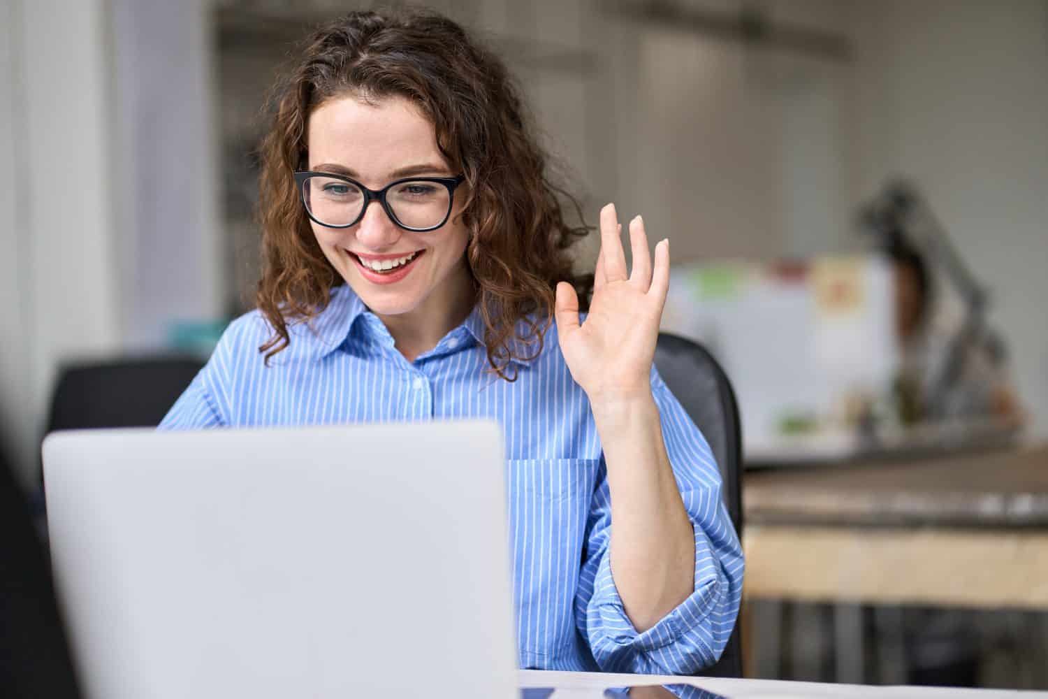 Young happy business woman using laptop waving hand greeting workers on remote virtual work conference meeting call in office. Hybrid video chat conversation corporate videoconference management.