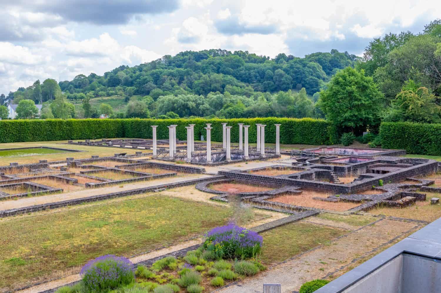 Restored ruins of roman villa in Echternach lake park, pillars, small stone walls, mountains with abundant green leafy trees in background, sunny summer day in Luxembourg