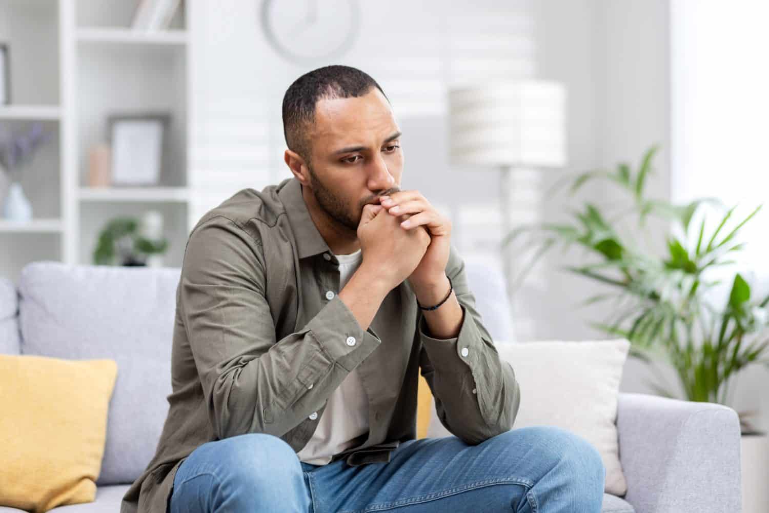 Depression in a young Latin American man sitting thoughtfully and upset at home on the couch.