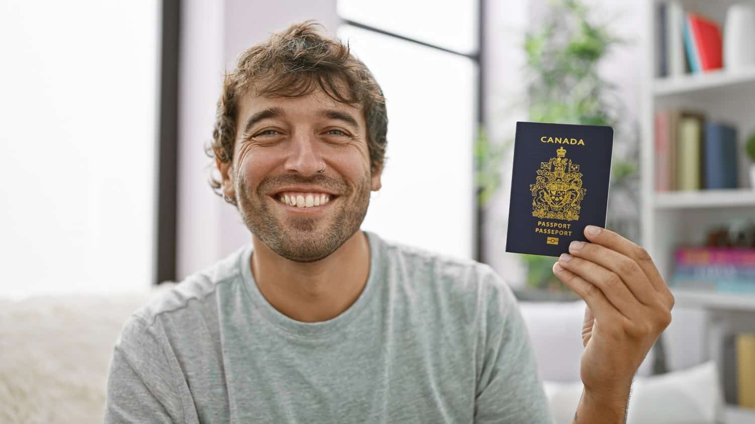 Happy young man, smiling and sitting at home on sofa, holding his canadian passport, ready for vacation!