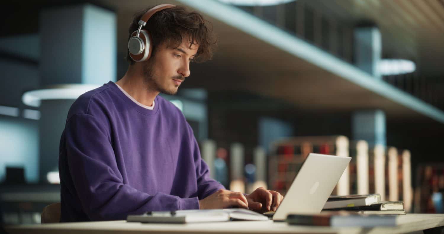 Portrait of a Smart Male Putting On Headphones and Working on a Laptop Computer. Young Man Doing a Homework Assignment and Preparing for Political Science Exams in a College Library