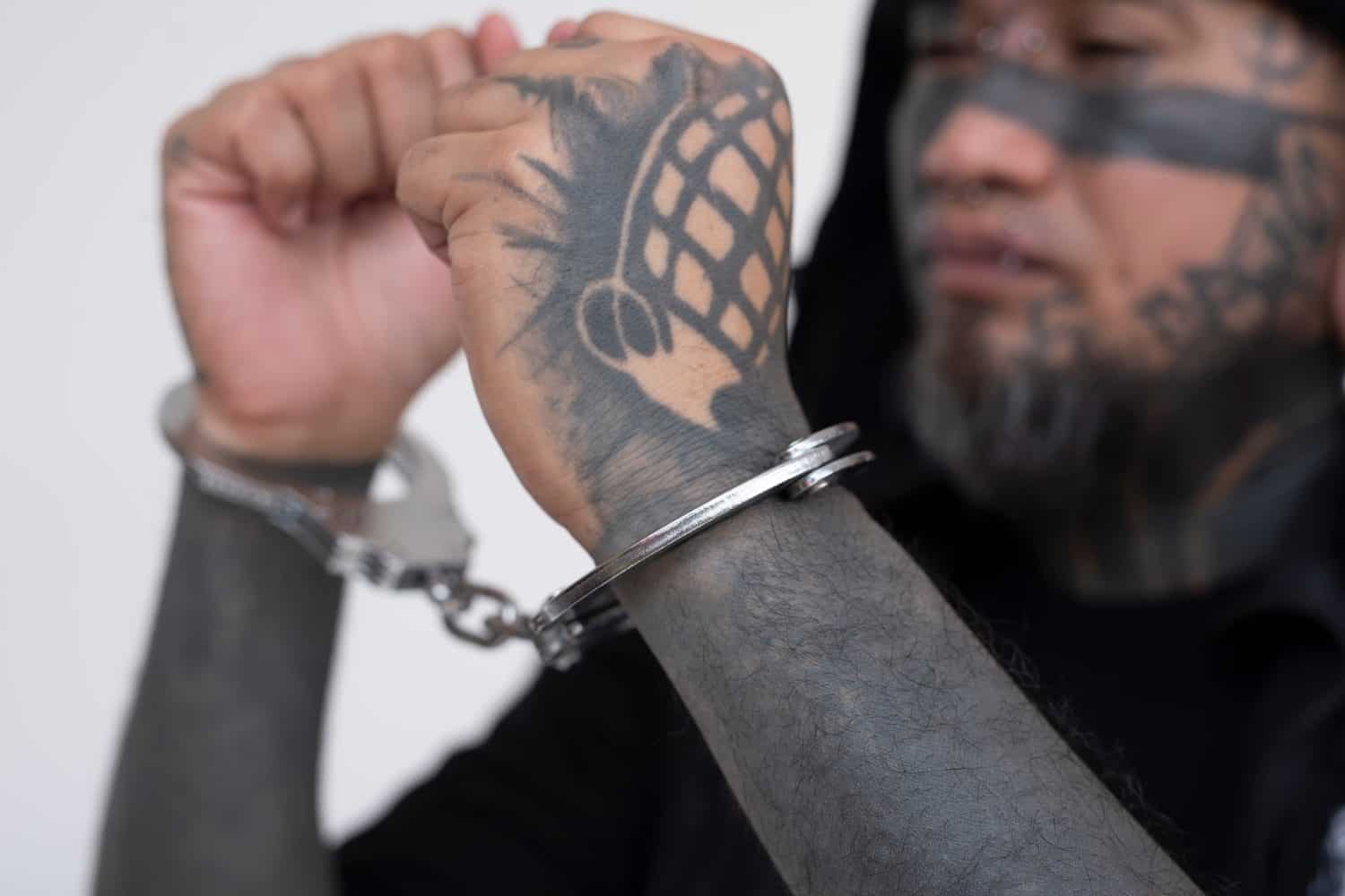 An Asian man with densely inked face tattoos, looking sideways in handcuffs, against a plain white background. A convicted felon under arrest. Judged and prejudiced by his looks.