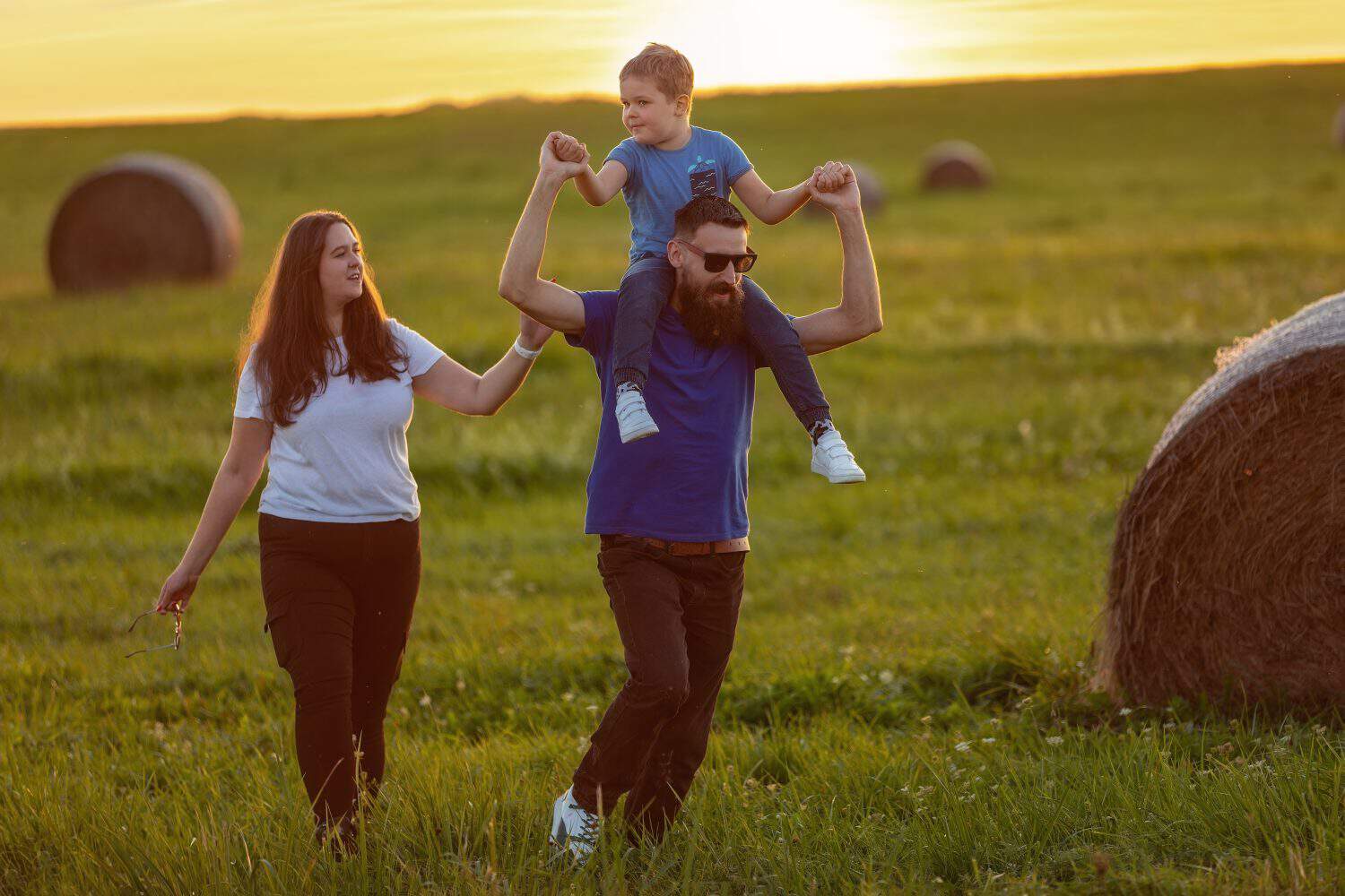 A young family with their little boy on their father&#039;s shoulders, running on a hilly meadow in the background of the sunset.