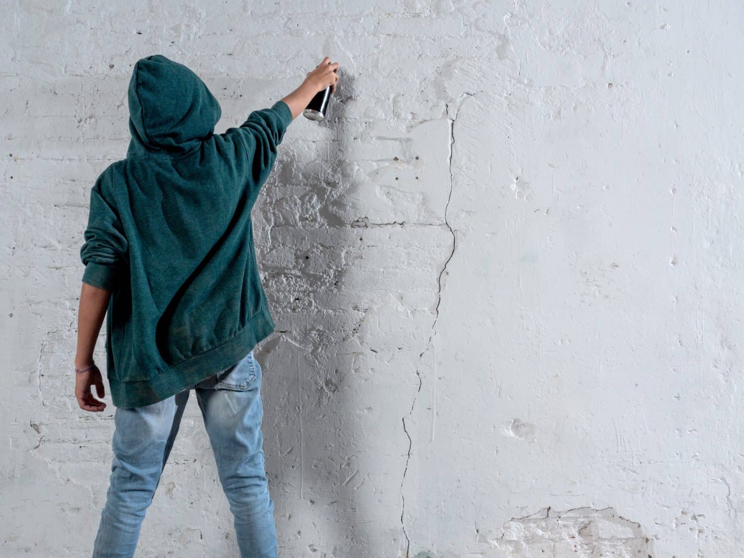 A young man in a green hoodie is spraying graffiti on a wall. Scene is rebellious and edgy, as the act of graffiti is often associated with vandalism and defiance of authority