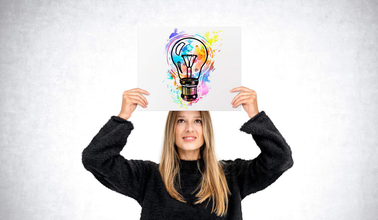 Smiling young woman showing poster with colorful lightbulb above head, grey concrete wall background. Concept of inspiration, start up, creativity, knowledge and idea