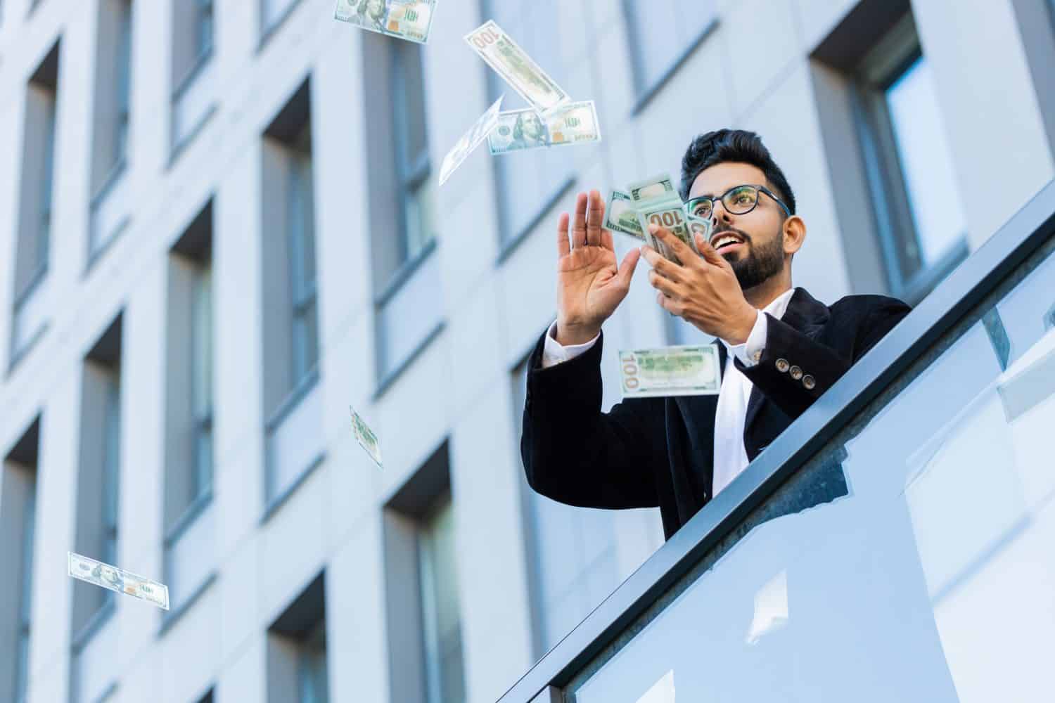 Happy rich Indian businessman wasting throwing money dollars cash around earnings, big profit, win lottery, share, celebrate, charity donation outdoors. Arabian Hindu freelancer guy in downtown city