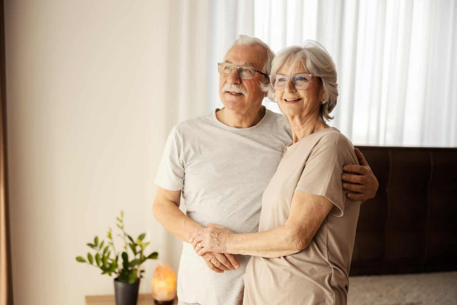 Smiling senior couple in love hugging at nursing home and looking through window.