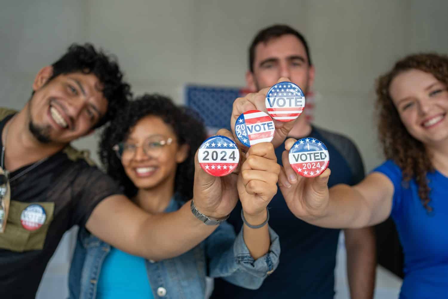 diverse group of smiling friends holding 2024 Vote election I voted buttons or pins at a US election polling station. In the background, american flag at the background, democracy in America USA