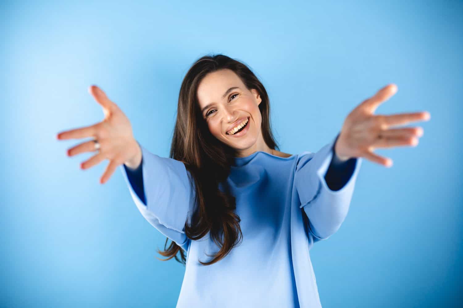 Cheerful lady making come to me sign. Pretty young girl with brunette hair look to camera, smiling and wear blue long sleeves top. Woman invite someone or want hugging, open hands, come to me.