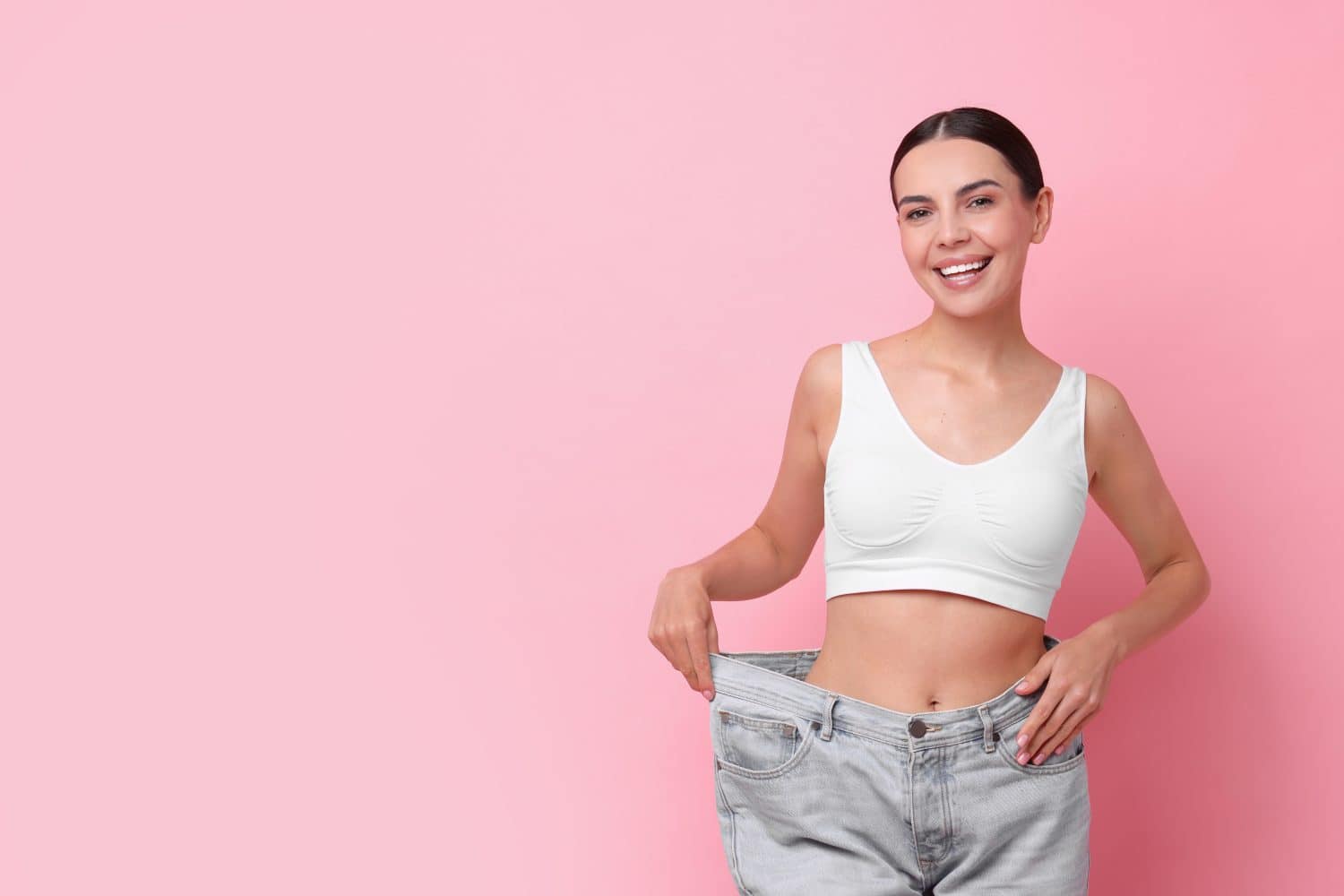 Diet and weight loss concept. Happy young woman in big jeans showing her slim body on pink background. Space for text