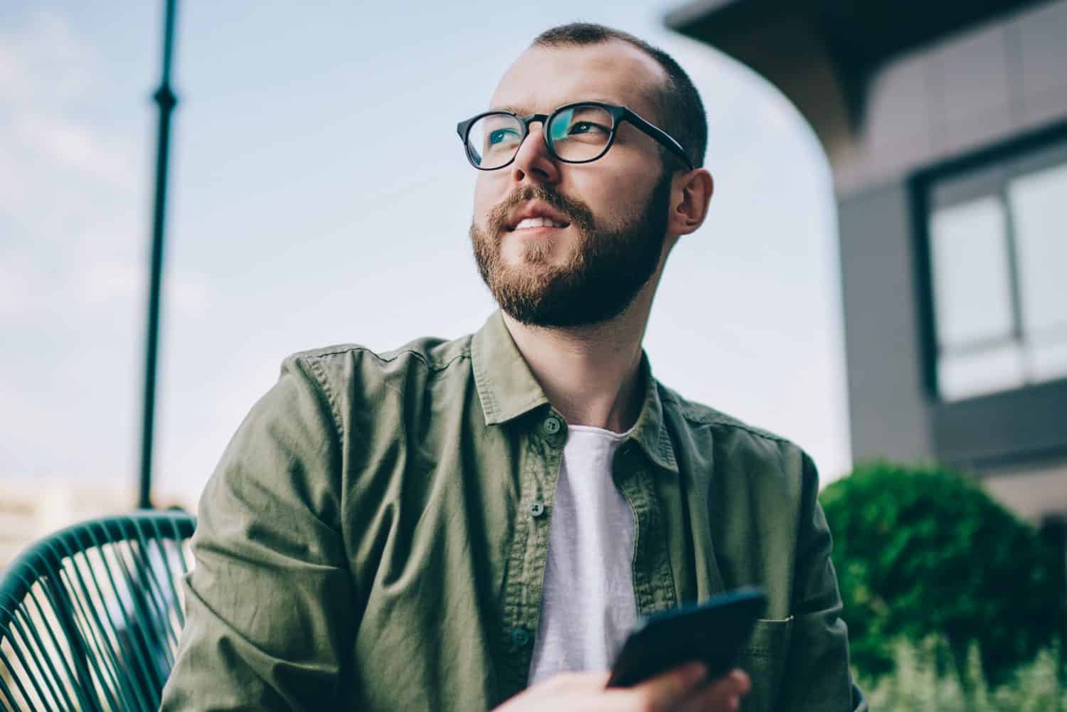 Pondering bearded young man looking away while thinking on idea for publication in blog on smartphone.Thoughtful hipster guy updating profile on telephone device using high speed internet connection