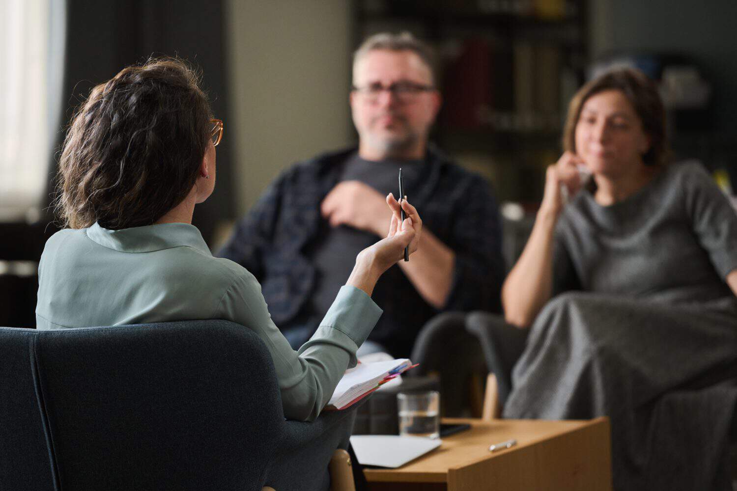 Couple having therapy session with female therapist gesturing while holding pen, room with bookshelf and therapy materials, focus on conversation, atmosphere calm and professional