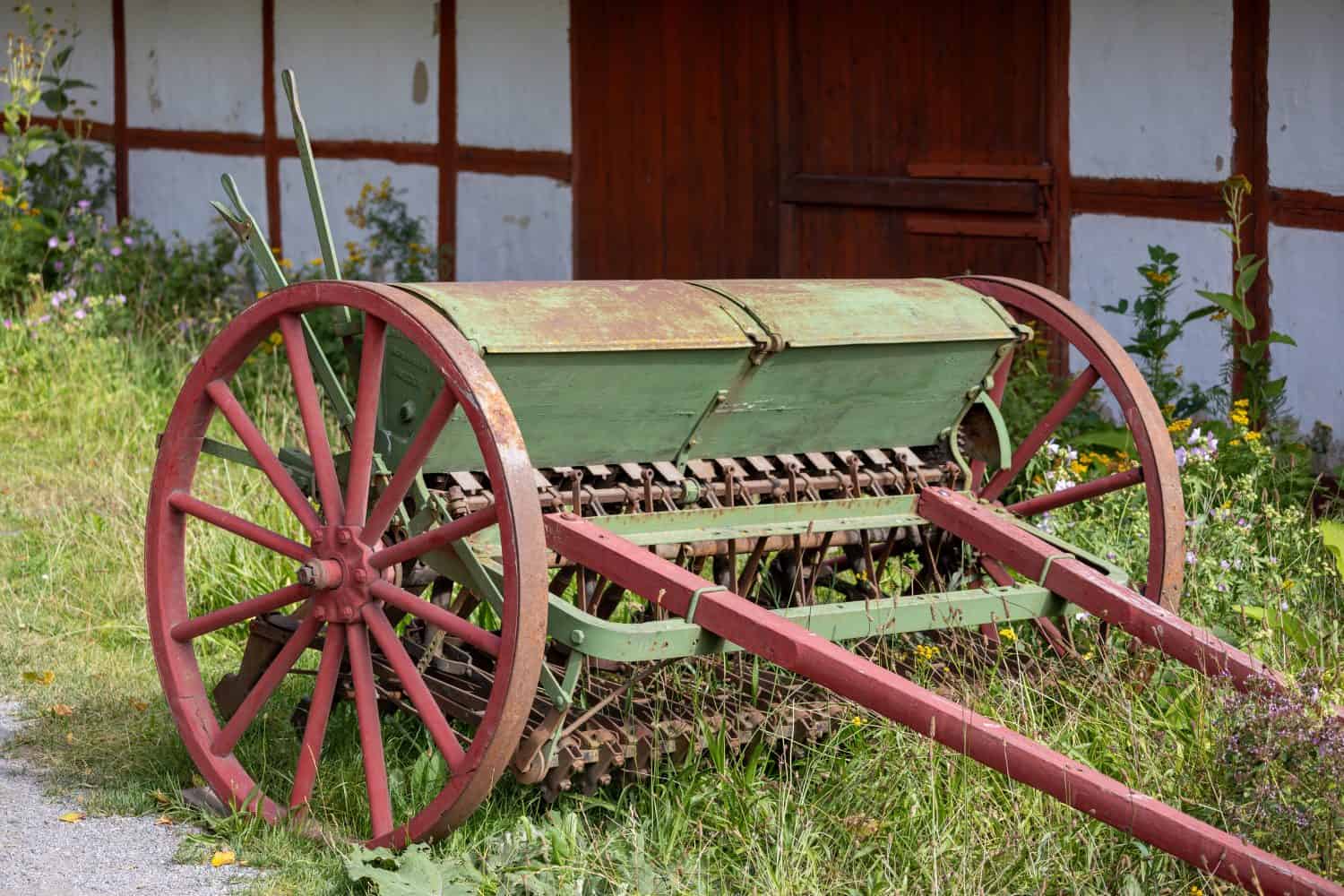 An old-fashioned green and red seed drill resting on grass, with a rustic wooden building in the background. The seed drill features large wooden wheels and a metal mechanism for planting seeds.