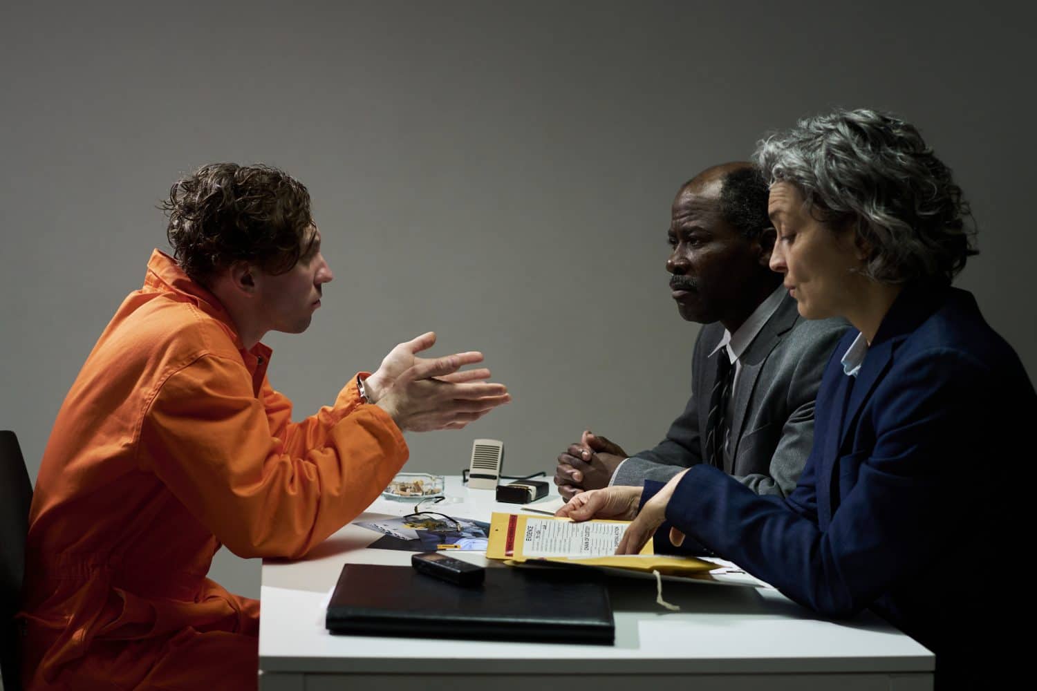 Inmate in orange uniform consulting with two legal advisors at meeting table, discussing important documents and strategies for upcoming court case