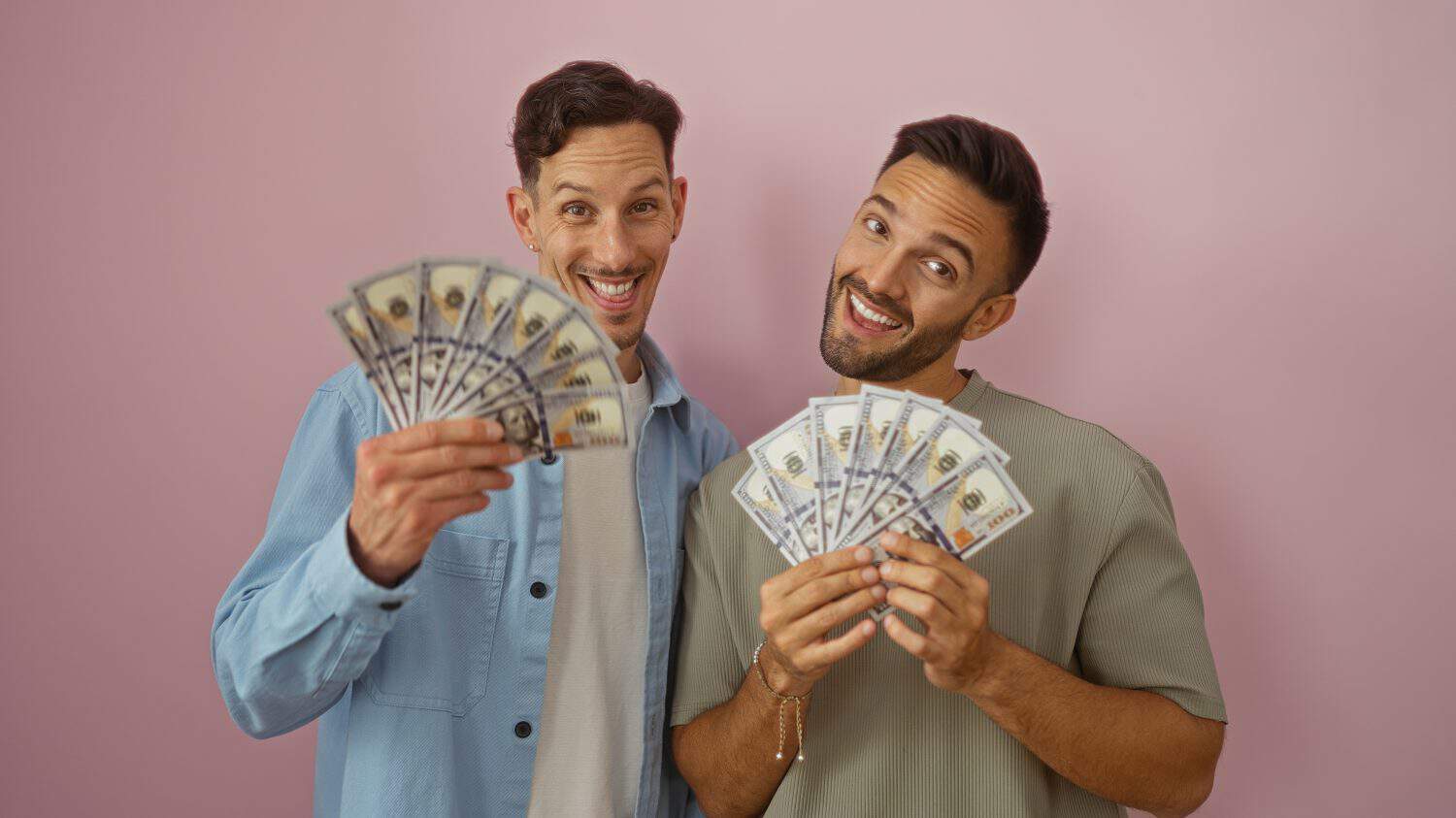 Couple, gay, lgbtq, men holding a spread of us dollars, smiling, and posing together against a pink background, symbolizing love, wealth, and happiness.