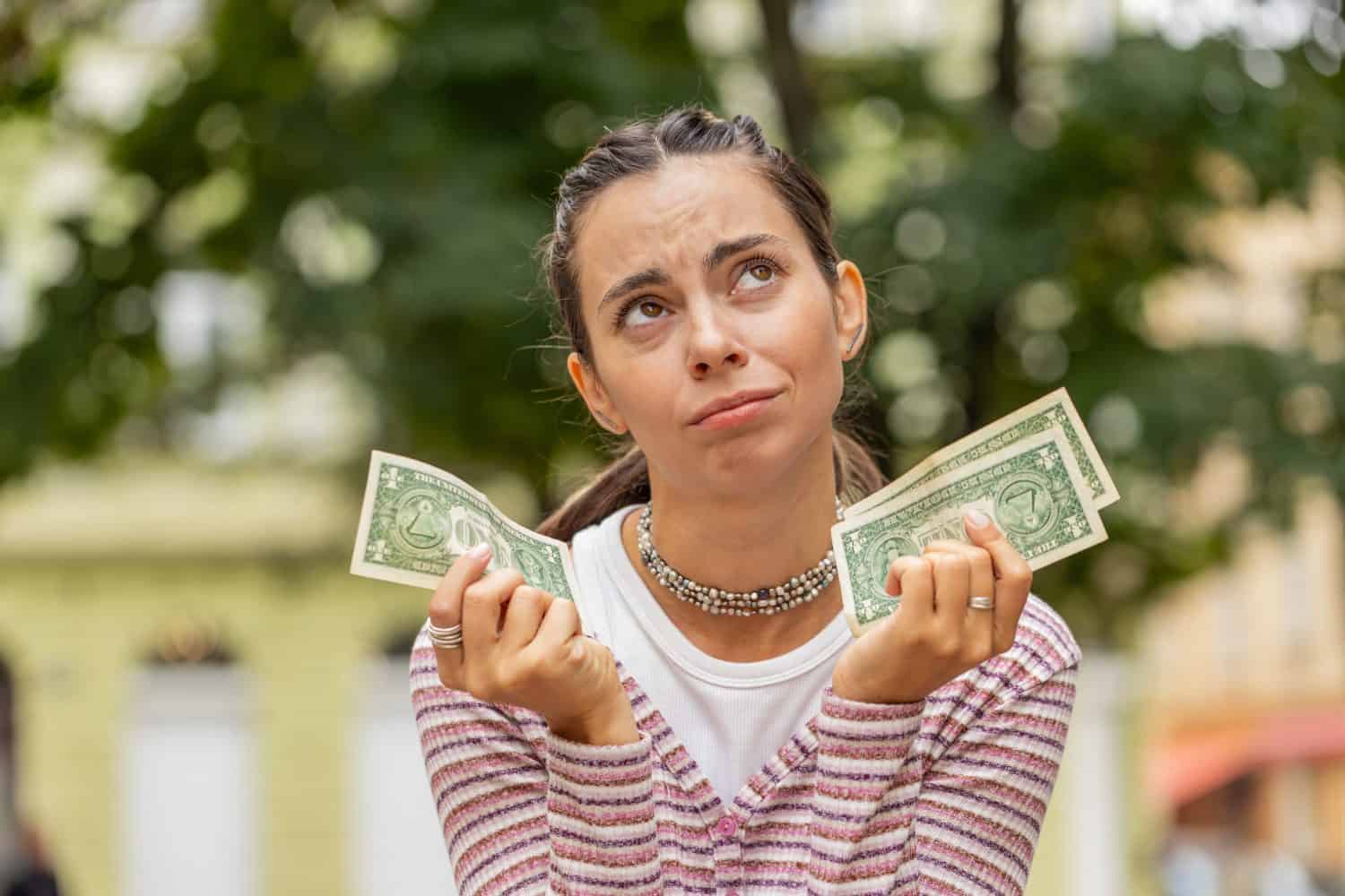 Poor Caucasian young woman holding one dollar banknotes outdoors. Lack of money to buy purchase something in store. Financial crisis. Bankruptcy. Poverty and destitution. Girl on urban city street