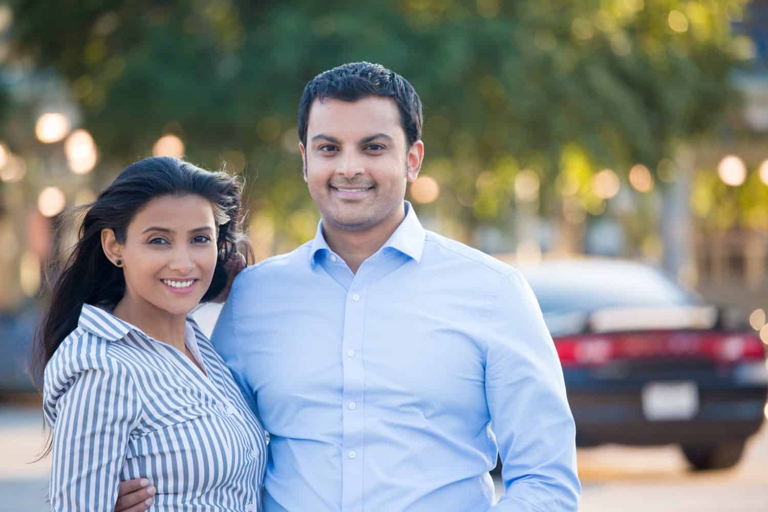 Closeup portrait, attractive wealthy successful couple in blue shirt and striped outfit holding each other smiling, isolated outside green trees and black car background.