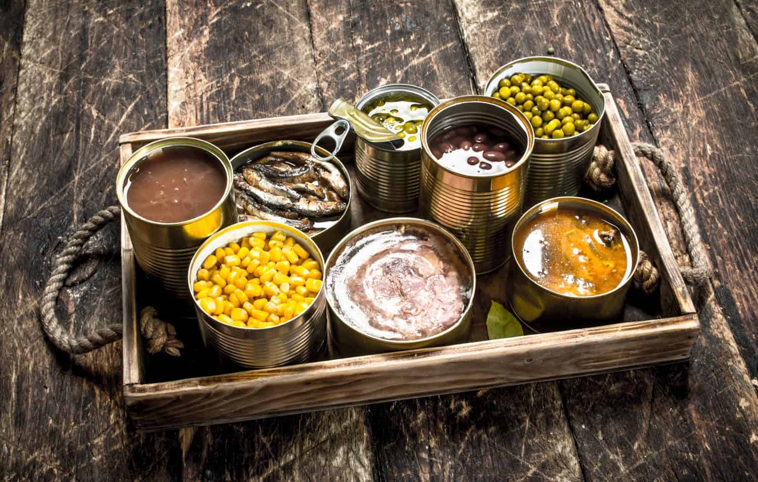 Various canned fruits, vegetables, fish and meat in tin cans on old tray. On a wooden background