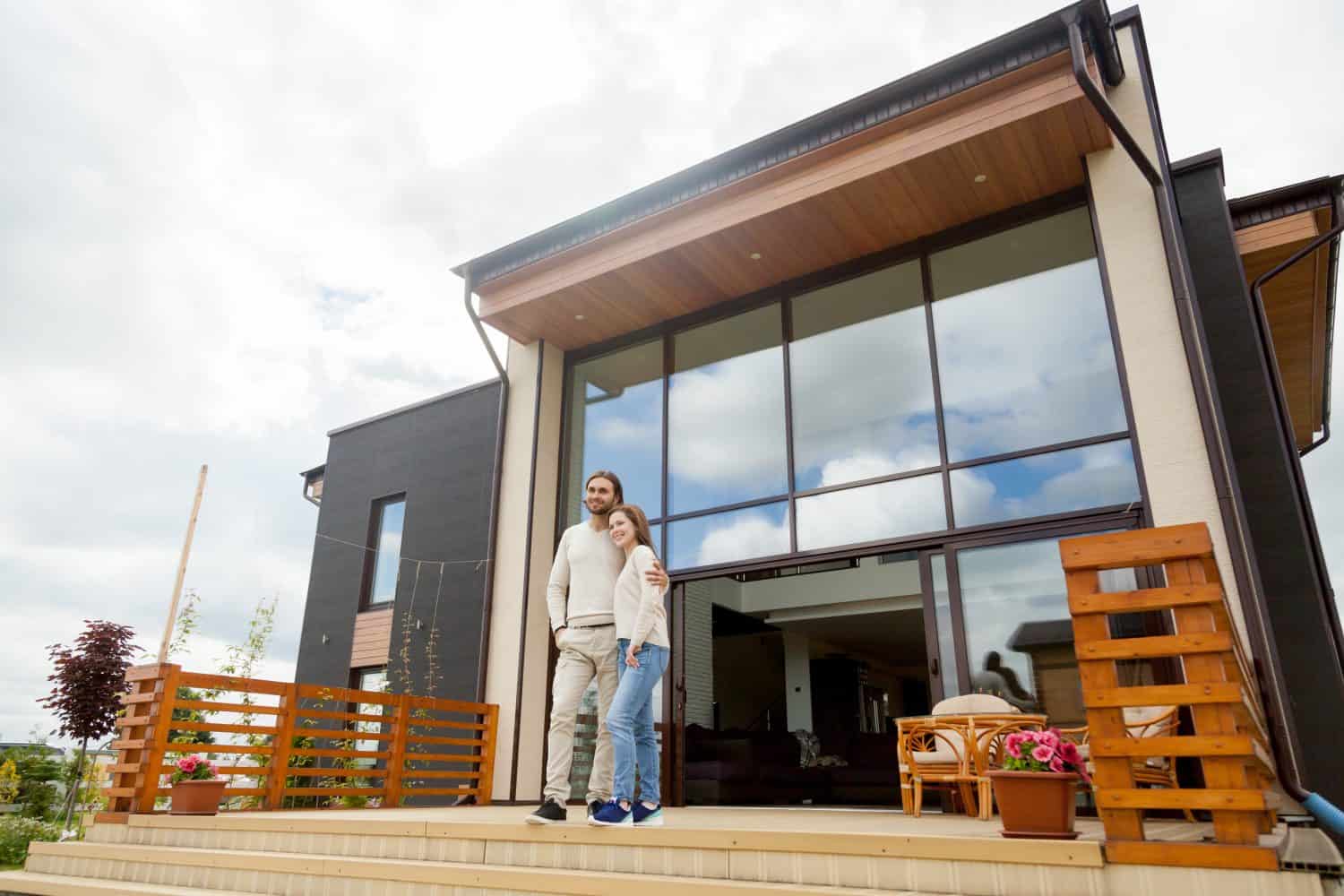 Happy young couple embracing standing outside big house on front porch, smiling owners enjoying luxury real estate wood and glass exterior design, buying home, mortgage loan, vacation rentals concept