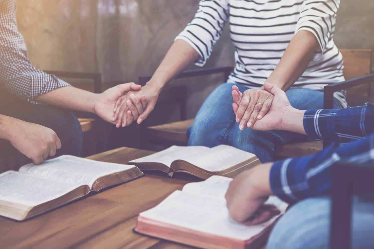 Christian small group holding hands and praying together around a wooden table with blurred open bible page in homeroom, devotional or prayer meeting concept