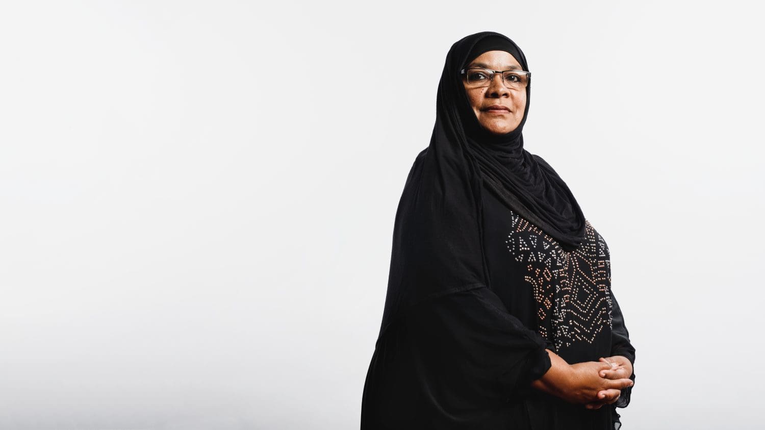 Senior muslim woman in hijab standing against white background. Arabic woman wearing eyeglasses and a black hijab looking at camera.