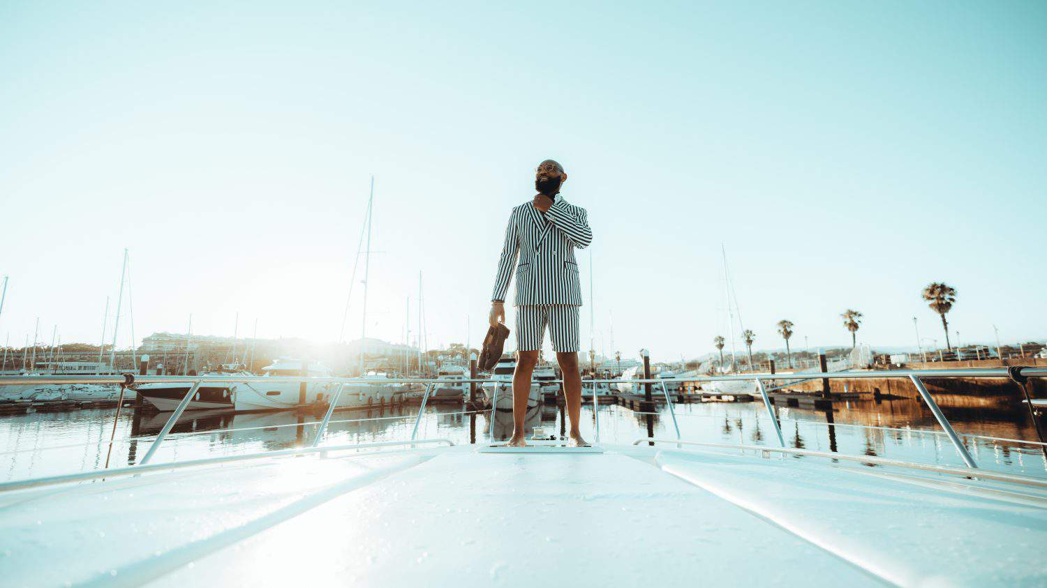 Wide-angle shot of a happy wealthy black man in a striped summer suit with shorts, standing on the bow of his luxury white yacht moored near the pier on a warm sunny evening, holding his shoes in hand