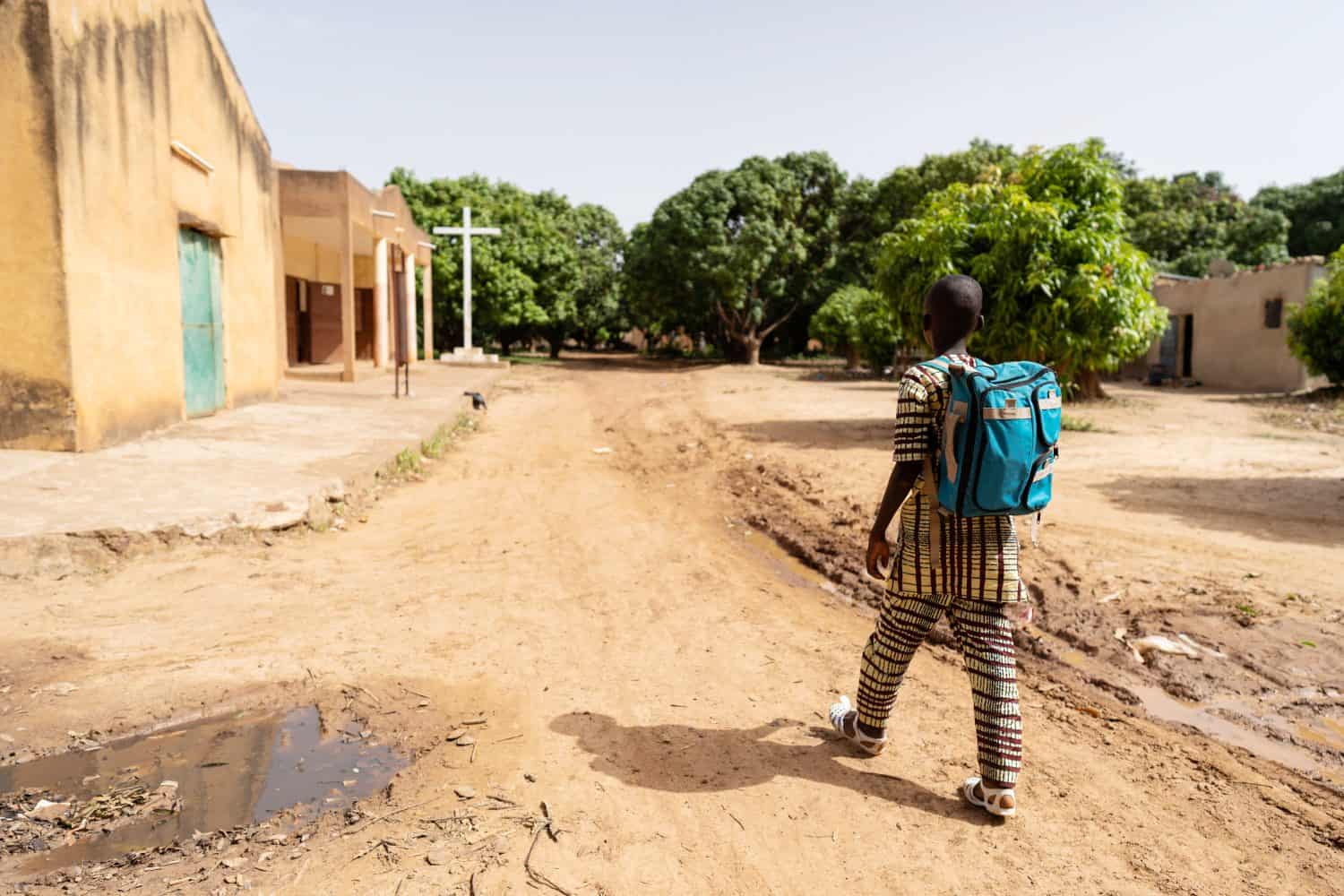 In this image, a lonely well dressed black boy with blue backpack on the way to sunday school or mass in a deserted Christian village in Africa