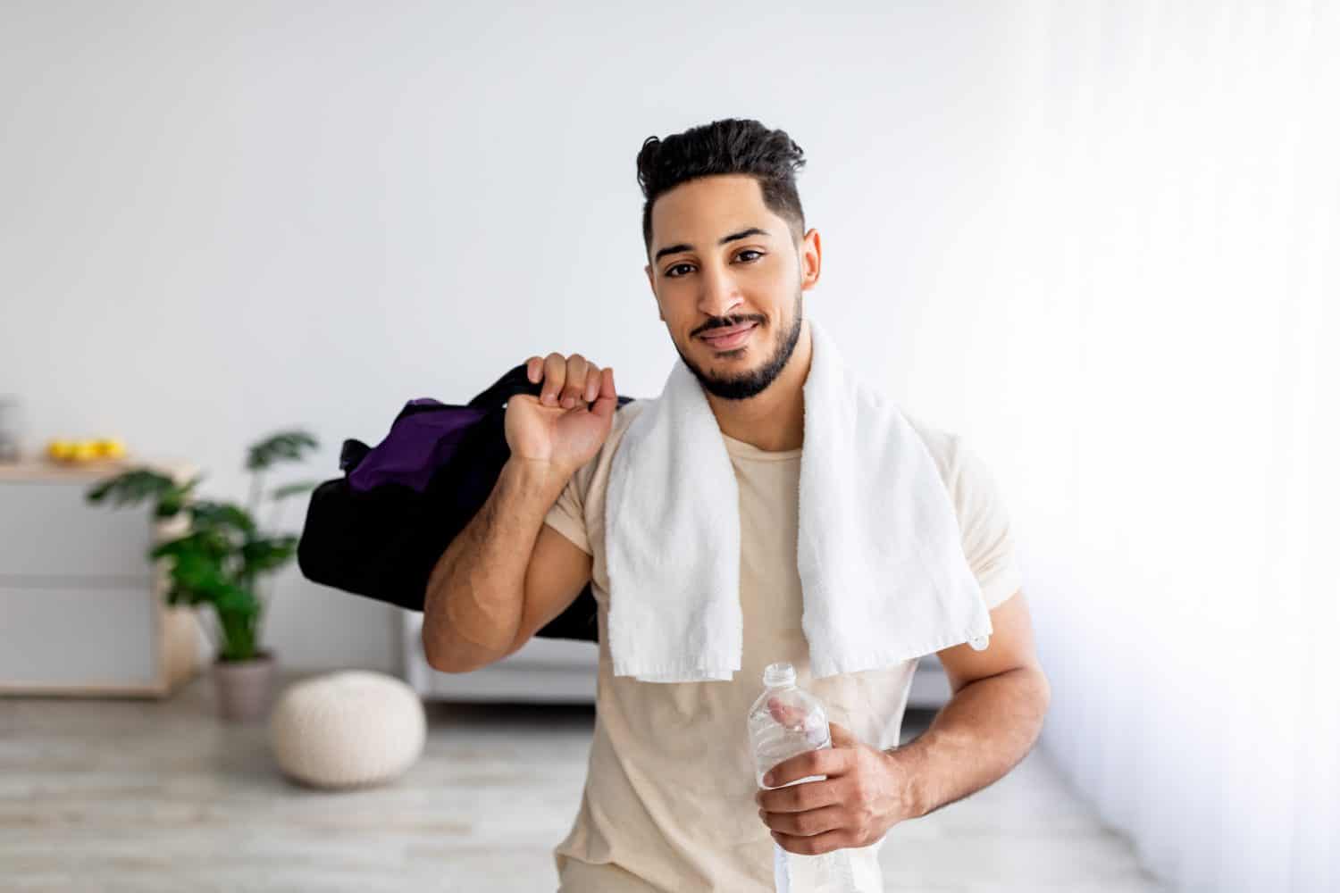 Handsome young Arab guy holding bottle of water and sports bag, posing after domestic training indoors. Attractive millennial Eastern man keeping hydrated during home workout