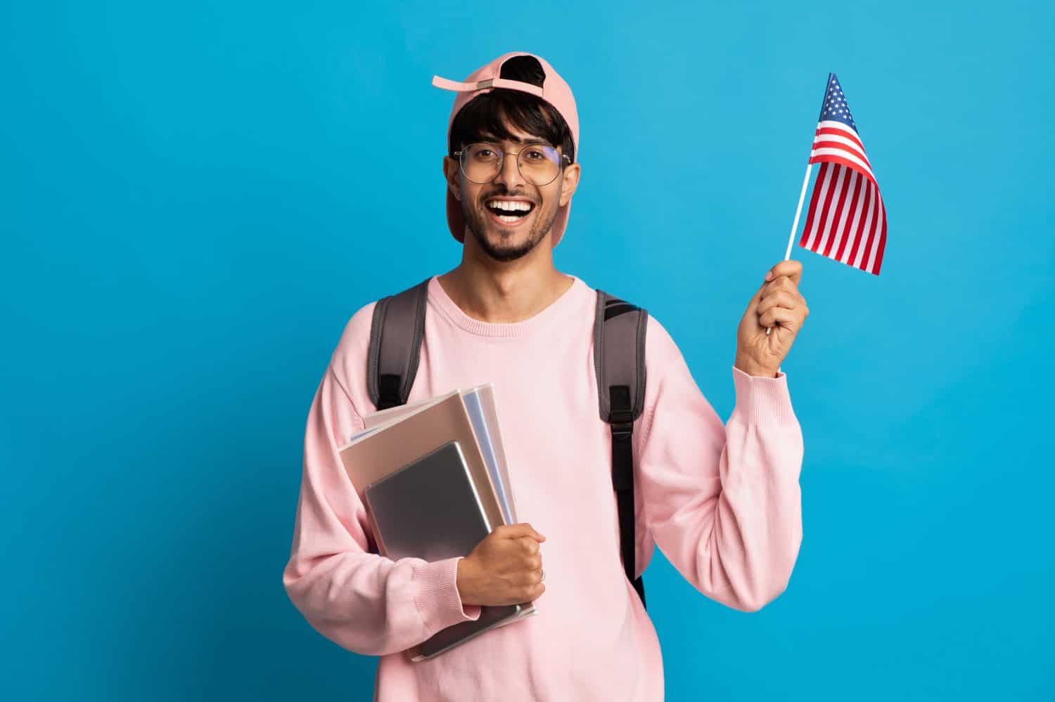 Smiling indian guy student with backpack and bunch of books showing flag of the US over blue studio background, copy space. Positive middle eastern young man studying English at school