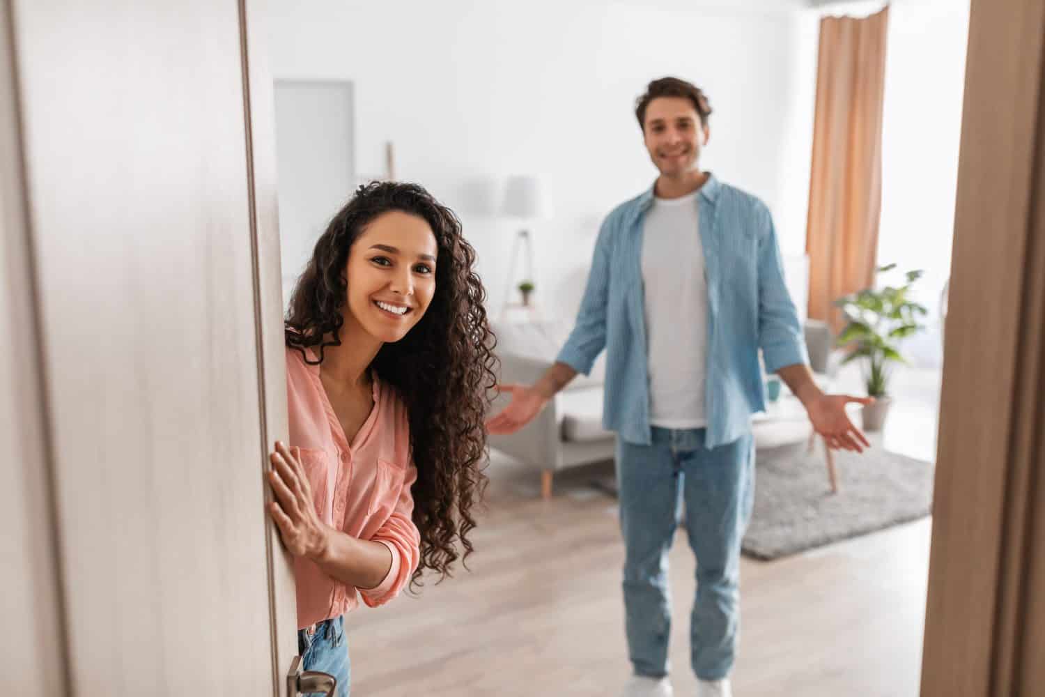 Portrait of cheerful couple inviting guests to enter home, happy people standing in doorway of modern flat, looking out waiting for visitor to come in, receiving friend, lady peeking out of front door