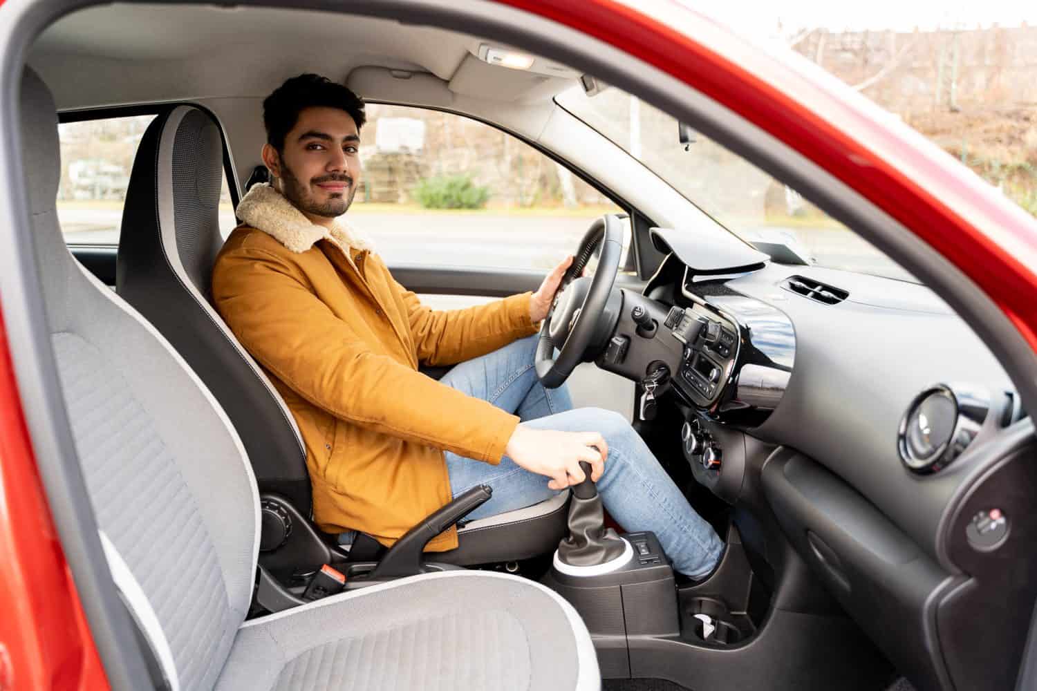 Portrait of spanish or arab eastern ethnicity man in yellow jacket holding driving wheel and manual gear box in car on sunny autumn, winter or spring day. Travel, exam, lesson, learning, taxi driver