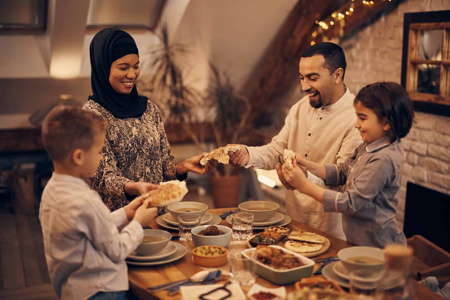 Happy Muslim parents and their kids sharing pita bread while eating dinner on Ramadan at home.