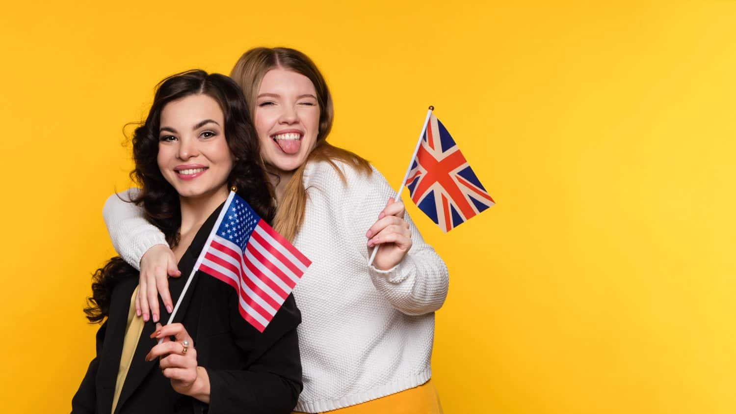 Two Girlfriends holding flags UK and USA. Study Abroad Concept. International Student Exchange Program. Young Women Hug Embrace, Have Positive Emotions, and Looking at Camera on Yellow Background.