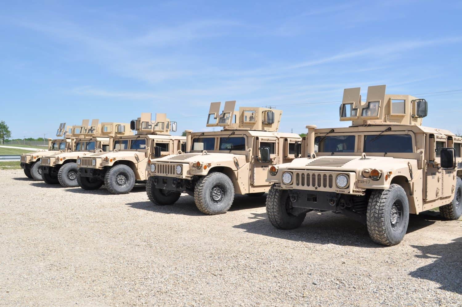 Desert tan US Military armored High Mobility Multi-Wheeled Vehicles (HMMWV) often used in the wars in Afghanistan and Iraq parked in a gravel lot with a blue sky and green grass in the background.