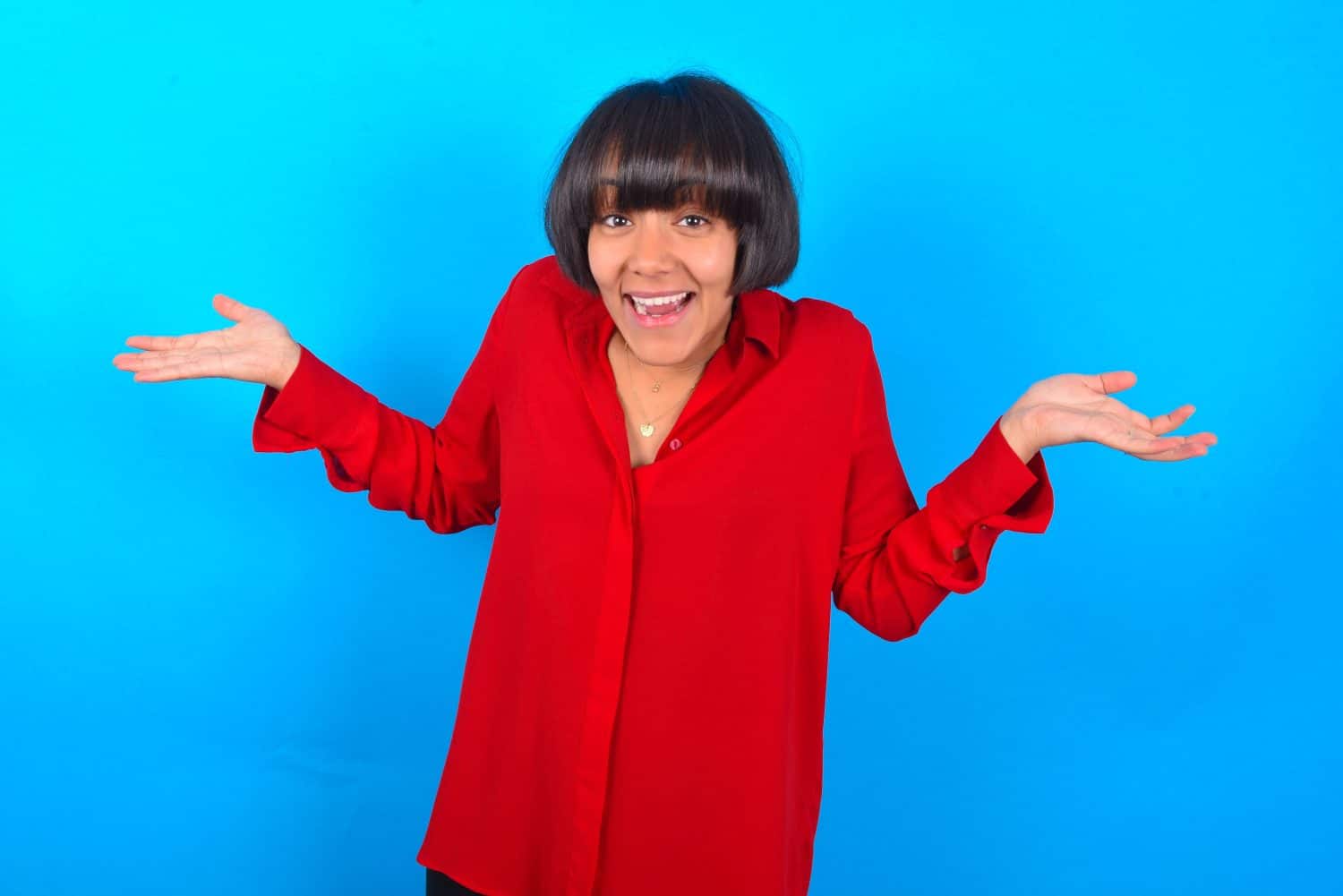 So what? Portrait of arrogant Young woman with bob haircut wearing red shirt over blue wall shrugging hands sideways smiling gasping indifferent, telling something obvious.