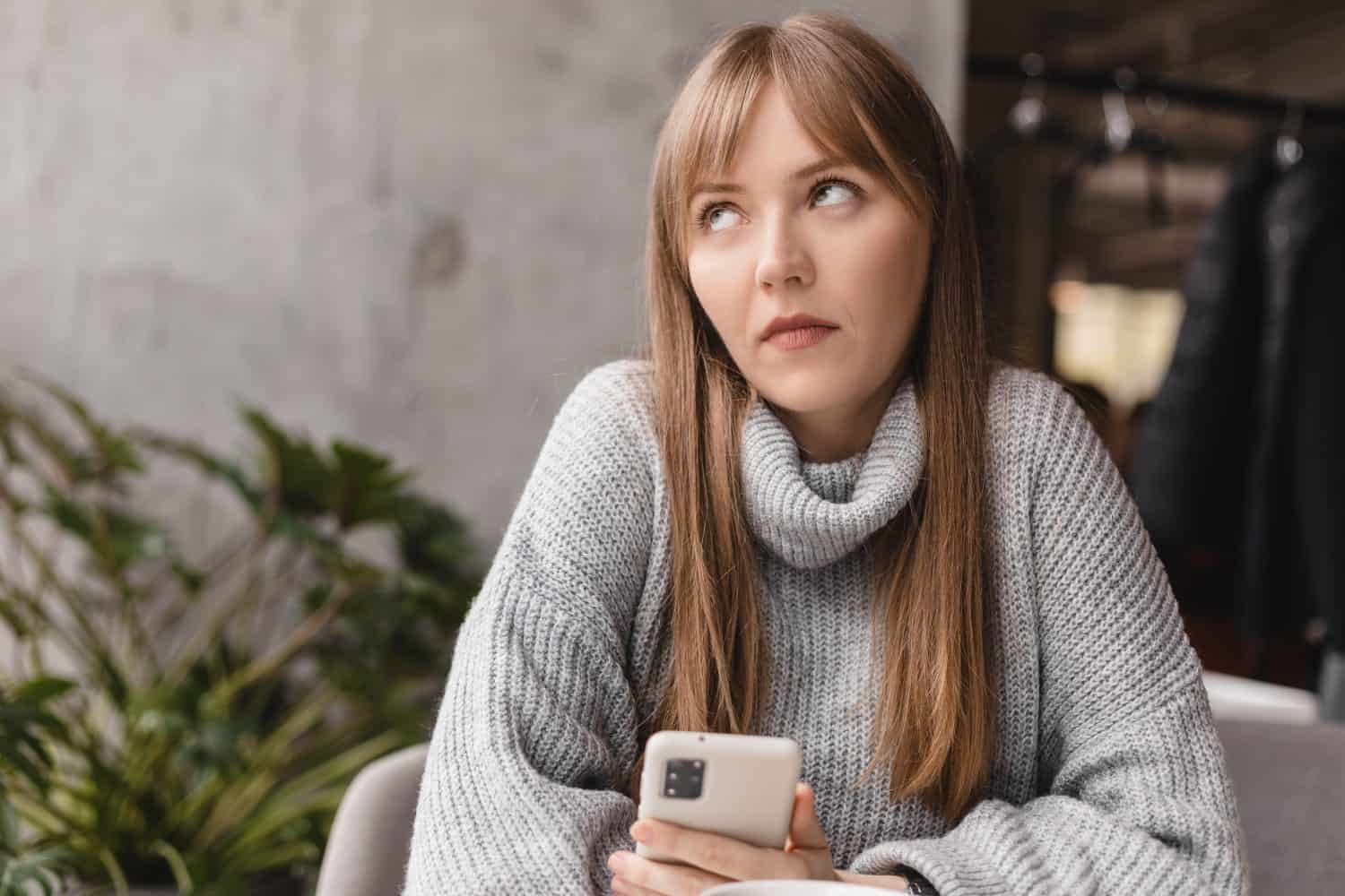 Confused puzzled blonde bang woman in casual clothes, sits in cafe, holds a smartphone in her hand, looks questioningly, rolling eyes. Girl getting surprising bad news, annoyed woman.