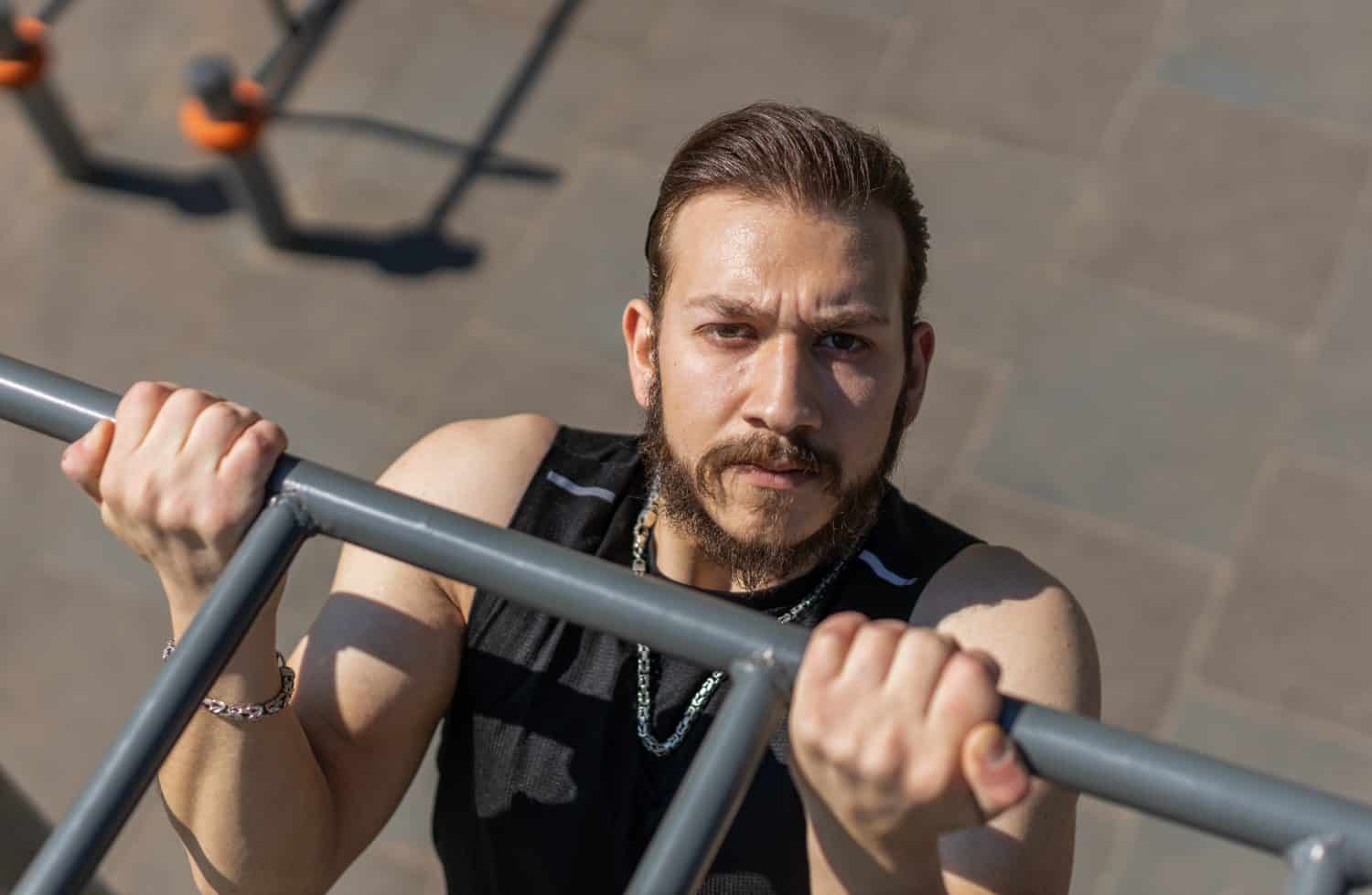 Athletic lebanese man in sportswear doing pull ups exercises on horizontal bar. Young guy pumping up back muscles on summer playground. Sports, health, fitness routine, workout. Strength, motivation