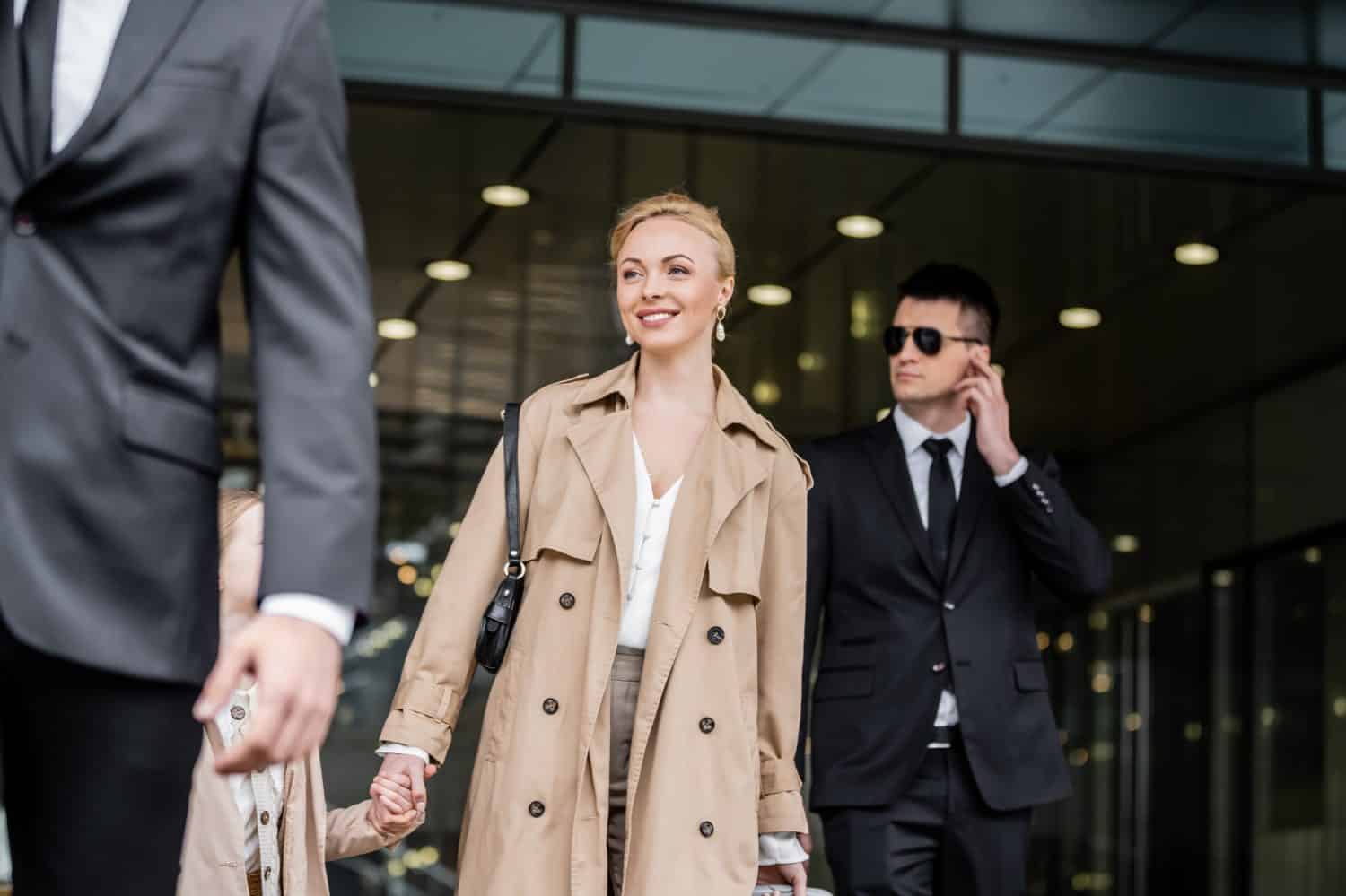 professional bodyguards protecting smiling blonde woman and preteen kid, successful mother and daughter in trench coats standing near hotel, safety and protection, private security, guards