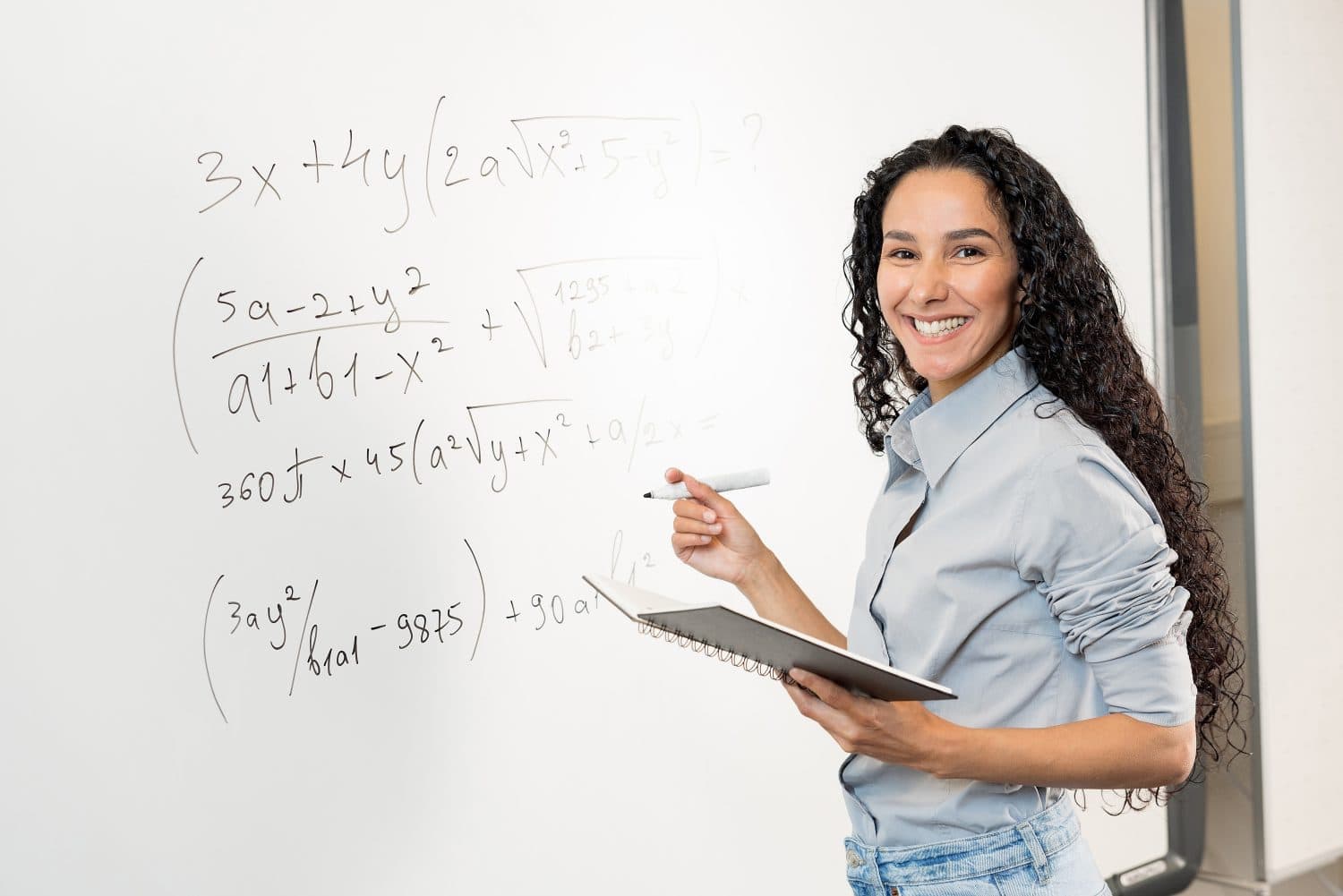 Smiling young business woman wearing a shirt looking at camera standing in classroom. Curly Arab teacher or professor posing for portrait in front of whiteboard. Holding a notebook, marker