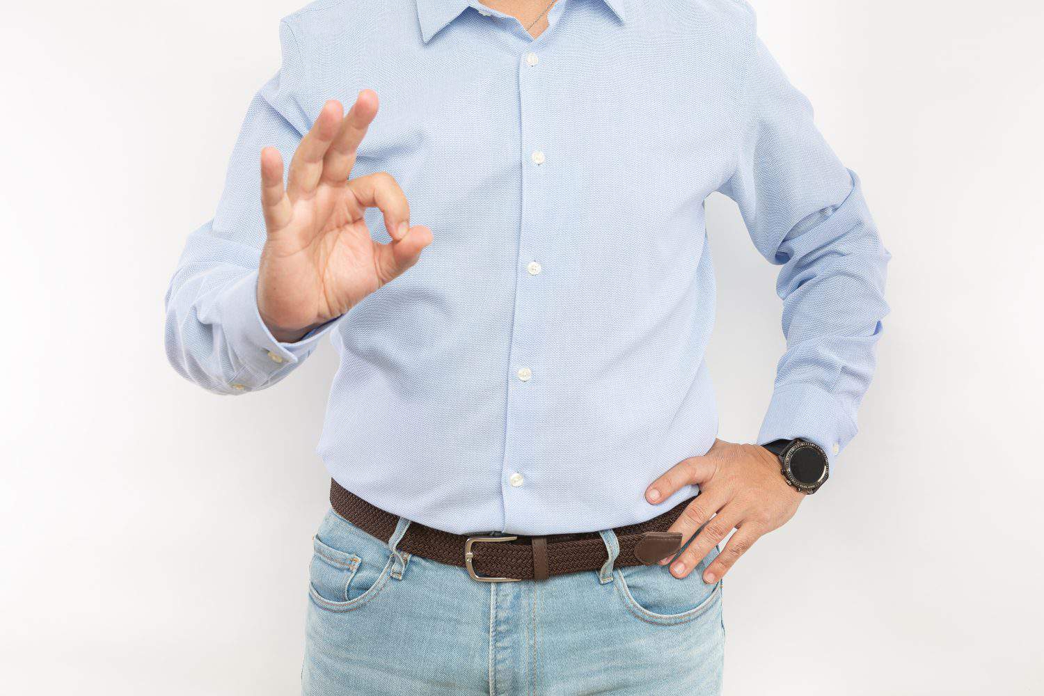 Crop unrecognizable male worker in shirt and jeans with smart wristwatch standing on white background while showing okay gesture