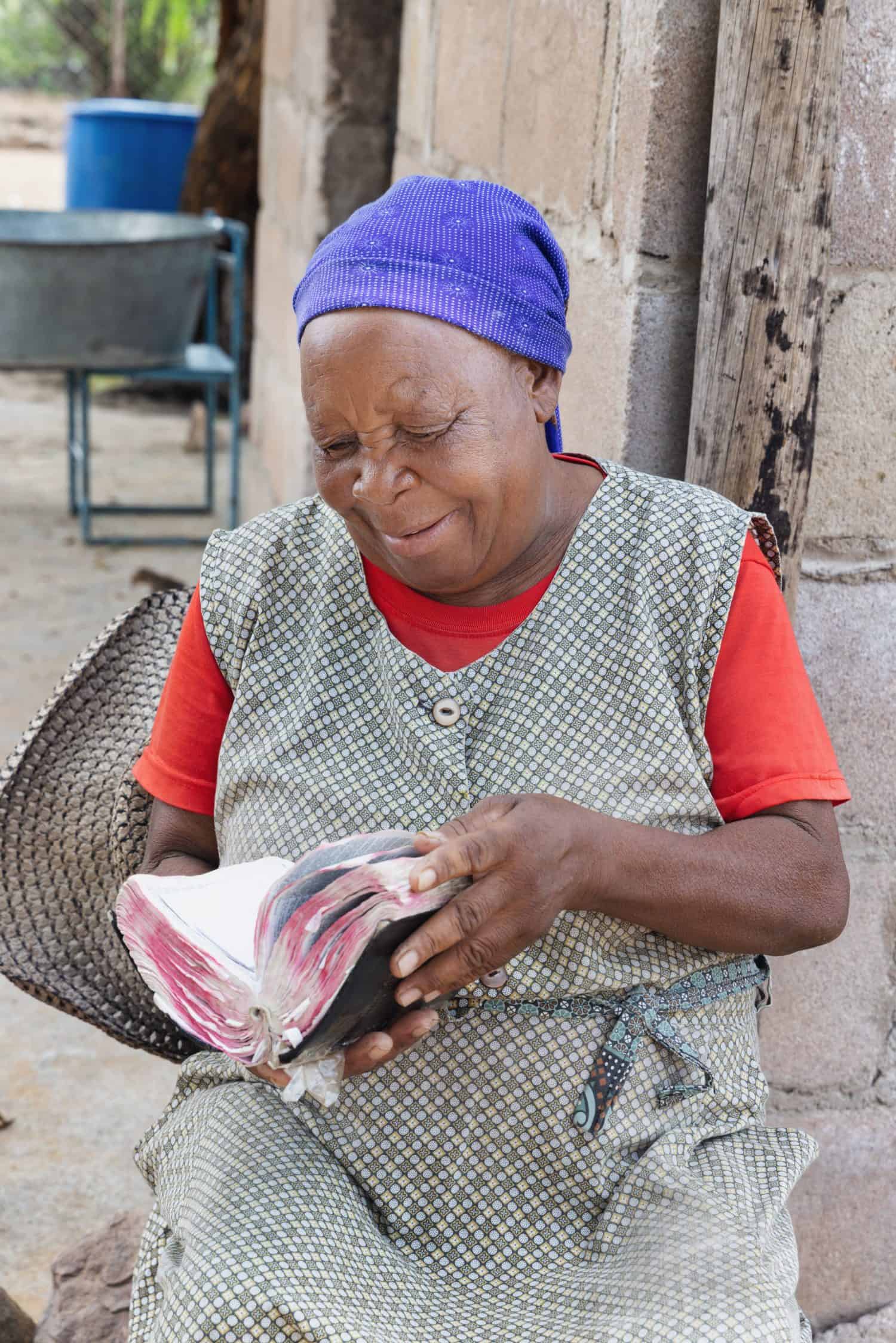 happy village religious Christian african old woman reading the bible, sitting in her yard, shack in the background